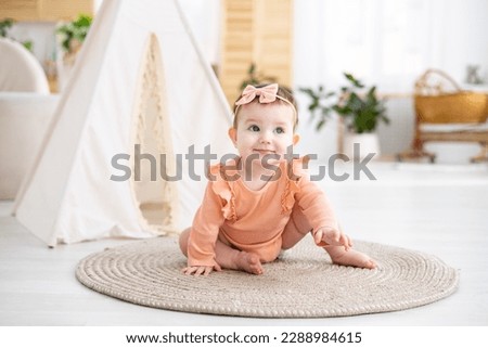 a cute little girl child in a pink bodysuit crawls on a rug in the living room of the house against the background of a wigwam, looks at the camera, smiles