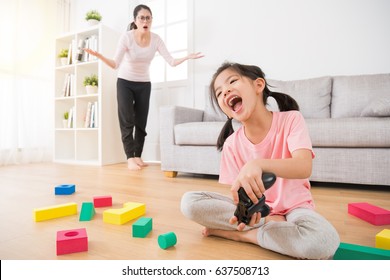 cute little girl children with a lot of messy toys on wood floor happy holding joystick play video games when her mother found stunned looking at her and make incredible gestures in background.