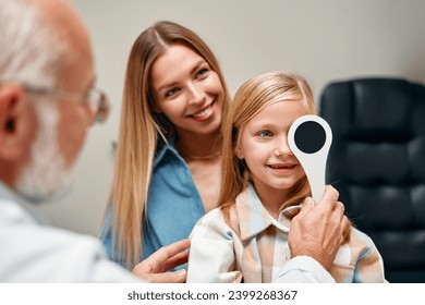 Cute little girl closing one eye during an ophthalmologist consultation while sitting on her mother's lap. A child points to an eye chart during an eye test. - Powered by Shutterstock