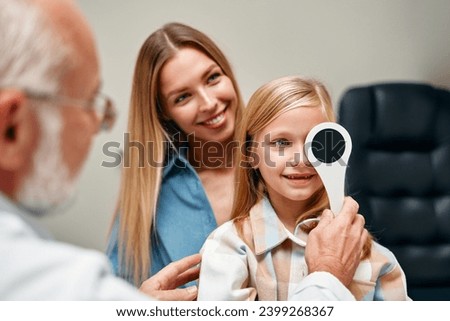 Cute little girl closing one eye during an ophthalmologist consultation while sitting on her mother's lap. A child points to an eye chart during an eye test.