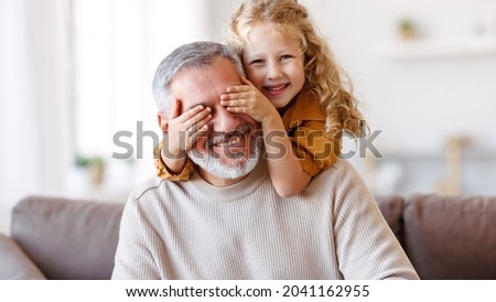 Cute little girl covering eyes with hands of her smiling senior grandfather while playing and having fun together at home, small happy child spending time with positive active grandpa in living room