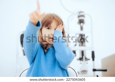 
Cute Little Girl Covering One Eye During Ophthalmological Consult. Toddler child pointing to a vision chart during eye check

