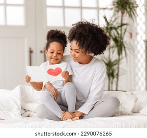Cute little girl daughter congratulating his mom happy african american woman and Mother's day  giving her handmade greeting postcard and red heart at home  Family holidays concept