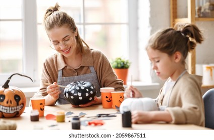Cute little girl daughter holding paintbrush   while painting Halloween pumpkins together at home  happy family mother   kid preparing handmade decorations for Saints Day party