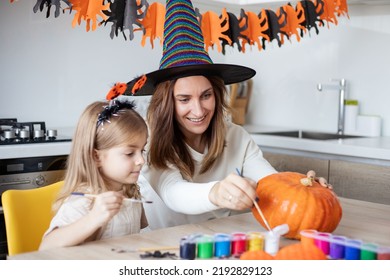 Cute little girl daughter holding paintbrush while painting Halloween pumpkins together at home
