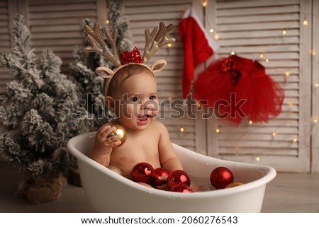 A cute little girl with deer horns on her head splashes in a bath with red and gold Christmas balls. The girl is happy