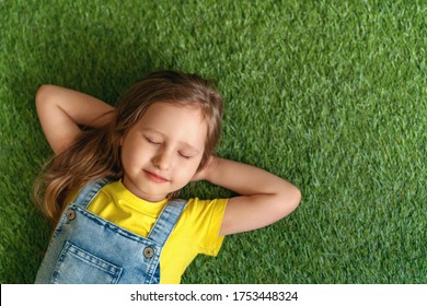 Cute Little Girl In Denim Overalls, Lying On A Green Carpet, Lawn. The Child Is Resting Lying On The Grass With His Eyes Closed. The Concept Of A Carefree Childhood, A Long-awaited Summer.