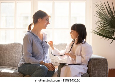 Cute Little Girl As Doctor Playing With Happy Mom Pretending Nurse, Playful Kid Daughter Dressed As Medical Worker Have Fun Holding Stethoscope Listening To Mother Chest, Funny Child Mum Game At Home