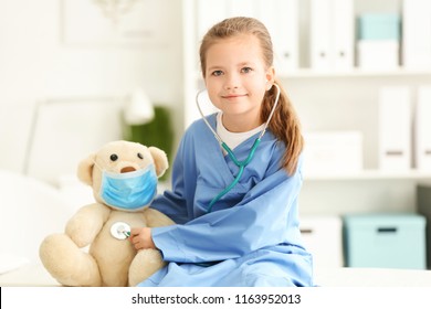 Cute little girl in doctor uniform playing with toy bear in hospital