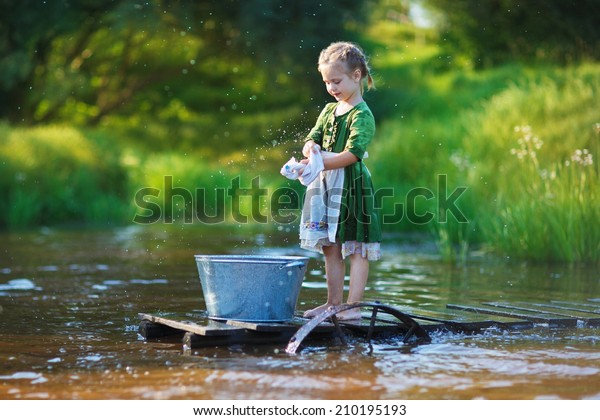 A cute little girl doing the laundry in a small\
aluminium basin standing on a pontoon at the pond in a sunny summer\
day. Kids are playing.
