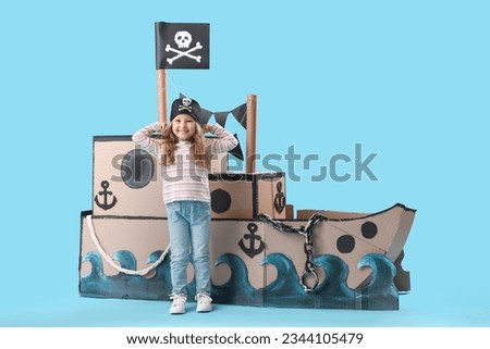Cute little girl dressed as pirate with cardboard ship on blue background