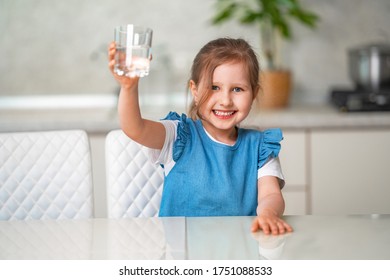 Cute Little Girl Drinking Water In The Kitchen At Home. The Little Girl Is Smiling Happily, Holding A Glass Of Clean Water And Wants To Drink It. Water Balance. Prevention Of Dehydration