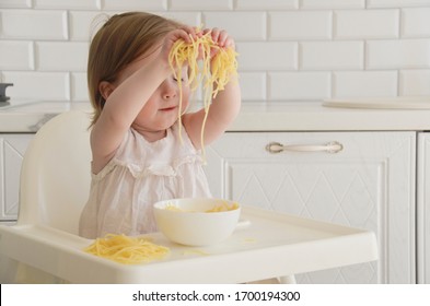 Cute Little Girl Eating Pasta? Baby Girl Sat In The White Kitchen. Baby Playing Food. Space For Text. Baby Girl Eat Corn Pasta. Gluten Free.