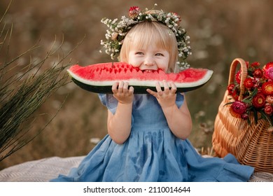 cute little girl eating big piece of watermelon on straw stack in summertime in the park. picnic with family. Adorable child wearing in flowers wreath on head and blue dress