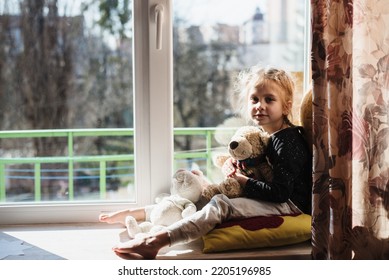 Cute Little Girl Enjoying The Sunshine While Sitting At The Window. The Child Is Playing Sitting On A Large Window Sill. Vitamin D. Film Noise