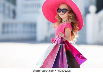 Cute Little Girl In Fashionable Hat On Shopping. Portrait Of A Kid With Shopping Bags. Child In Dress, Sunglasses And Shoes Near Shopping Mall Having Fun. Shopping. Girl. Fashion