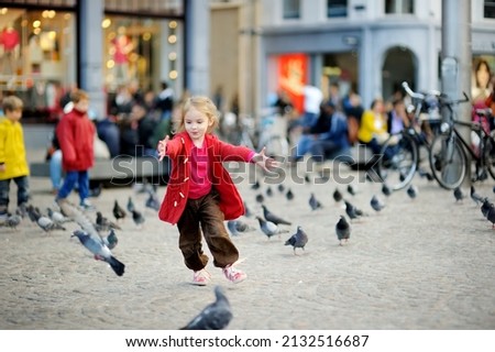 Cute little girl feeding and chasing birds on Dam Square in Amsterdam on summer day. Child feeding pigeons and sparrows outdoors.