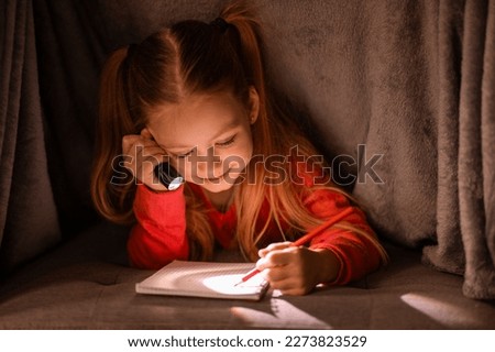 Cute little girl with flashlight hiding under blanket and writing in notebook, smiling preteen female child relaxing under covers and drawing in notepad, filling diary, using torch in the dark