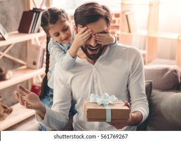 Cute little girl is giving her handsome father a gift box. Both are sitting on couch at home and smiling