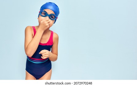 A cute little girl in goggles, a swimsuit, and a swimming cap holding her nose preparing for swimming isolated blue studio background. Kid in swimwear holding her nose to learning to swim in the pool. - Powered by Shutterstock