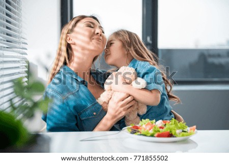 Cute little girl is going to kiss her mom with love. Asian woman closed eyes with happiness. They are sitting in kitchen and hugging