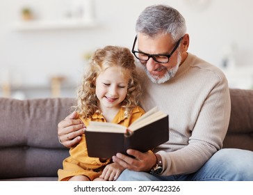 Cute little girl granddaughter reading book with positive senior grandfather while sitting on sofa together and enjoying leisure time on weekend at home  - Powered by Shutterstock