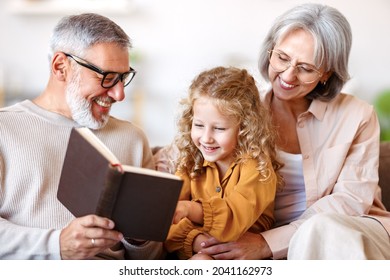 Cute Little Girl Granddaughter Smiling When Reading Book With Senior Grandparents While Sitting Together On Sofa In Living Room, Child Enjoying Leisure Time With Grandma And Grandpa On Weekend At Home