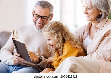 Cute little girl granddaughter smiling when reading book with senior grandparents while sitting together on sofa in living room, child enjoying leisure time with grandma and grandpa on weekend at home - Powered by Shutterstock