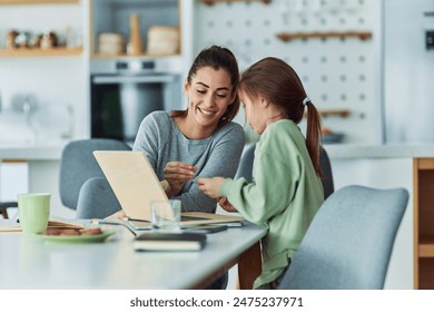 A cute little girl handing chalk to her mother as they study together at the dining table using a wooden laptop with a chalkboard screen. - Powered by Shutterstock