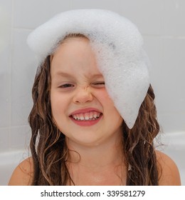 Cute Little Girl Having A Bath In Foam Water. Child Washing Her Long Hair. Girl Showing Her Tricky Silly Face. Close Up. Selective Focus On Kids Face.