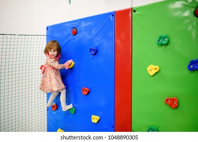 Cute Little Girl Having Fun In Leisure Center For Kids. Child Sport Activity In Indoor Playground. Toddler Girl Climbing On Small Mountaineering Wall