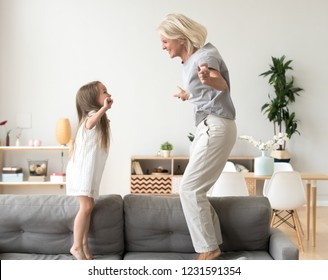 Cute little girl having fun playing with smiling grandmother jumping on couch together, happy granny and active kid grandchild dancing on sofa, grandma and granddaughter laughing playing at home