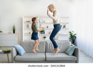 Cute Little Girl Having Fun Playing With Smiling Grandmother Jumping On Couch Together, Happy Granny And Active Kid Grandchild Dancing On Sofa, Grandma And Granddaughter Laughing Playing At Home