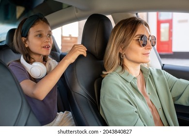 Cute Little Girl With Headphones Holding By Black Leather Seat Of Her Mom Driving Electric Car