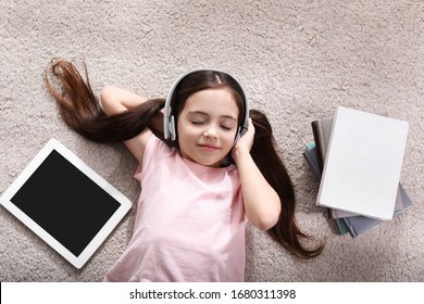 Cute little girl with headphones and tablet listening to audiobook on floor indoors, flat lay