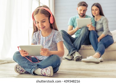 Cute little girl in headphones is using a tablet and smiling, in the background her parents are using a tablet too, sitting on sofa at home - Powered by Shutterstock