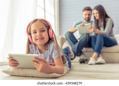 Cute little girl in headphones is using a tablet and smiling, in the background her parents are using a tablet too, sitting on sofa at home - Powered by Shutterstock