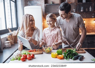 Cute little girl and her beautiful parents are using digital tablet and smiling while cooking in kitchen at home - Powered by Shutterstock