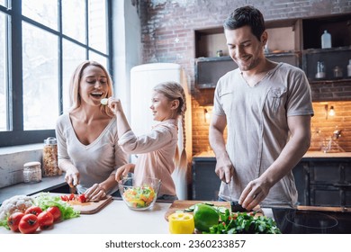 Cute little girl and her beautiful parents are cutting vegetables and smiling while cooking in kitchen at home - Powered by Shutterstock