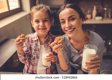 Cute little girl and her beautiful mother are looking at camera and smiling while drinking milk and eating muffins