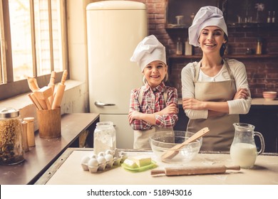 Cute little girl and her beautiful mother in chef hats are standing with crossed arms, looking at camera and smiling while baking at home