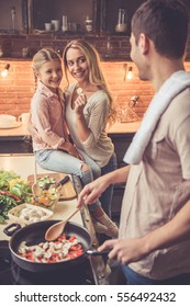 Cute Little Girl And Her Beautiful Mom Are  Smiling While Dad Is Cooking In Kitchen At Home