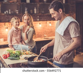 Cute Little Girl And Her Beautiful Mom Are  Smiling While Dad Is Cooking In Kitchen At Home