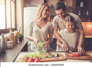 Cute little girl and her beautiful parents are cutting vegetables and smiling while making salad in kitchen at home