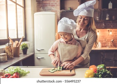 Cute Little Girl And Her Beautiful Mom In Chef's Hats Are Cutting Vegetables And Smiling While Cooking In Kitchen At Home