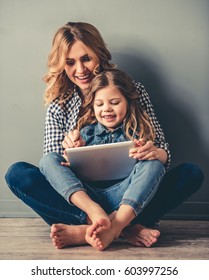 Cute Little Girl And Her Beautiful Young Mom Are Sitting Together On The Floor, Using A Digital Tablet And Smiling, On Gray Background