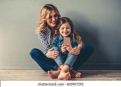 Cute Little Girl And Her Beautiful Young Mom Are Sitting Together On The Floor, Using A Smart Phone And Smiling, On Gray Background