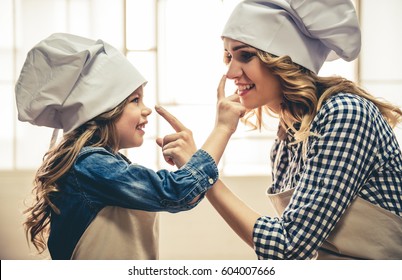 Cute Little Girl And Her Beautiful Mom In Aprons And Chef Hats Are Touching Noses And Smiling While Kneading The Dough In The Kitchen