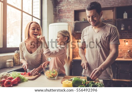 Cute little girl and her beautiful parents are cutting vegetables and smiling while cooking in kitchen at home