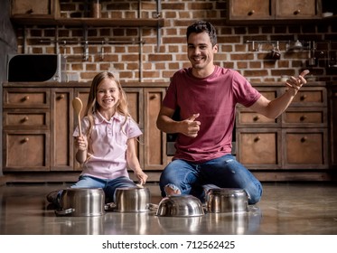 Cute Little Girl And Her Dad Are Using Wooden Spoons, Looking At Camera And Smiling While Playing Drums With Dishes In Kitchen, Dad Is Imitating Guitar